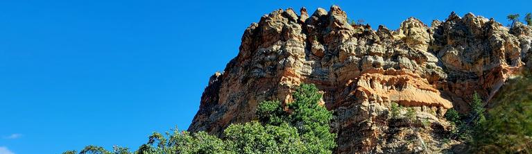 View of cliff and rock face in multiple colors with trees at the base and clear blue sky